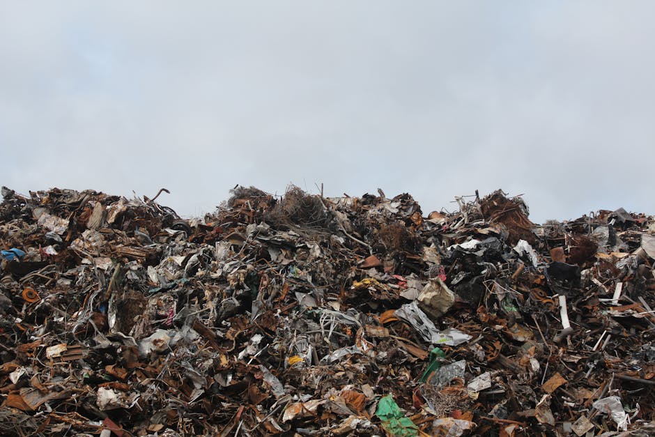 A large pile of mixed waste materials is shown in the foreground, consisting of fragmented and crumpled plastics, metal scraps, and broken electronic components. The waste appears to be discarded rubbish collected from various sources, with textures ranging from smooth, shiny plastic to rough, rusted metal surfaces. The colours in the pile include shades of black, grey, brown, green, and hints of orange and blue, reflecting different materials and contamination. The waste is loosely stacked, creating an uneven, jagged surface that dominates the lower half of the image. In the background, a cloudy sky dominates the upper portion of the image, providing a neutral, overcast atmosphere. The scene appears to be outdoors, possibly on an open site or waste management area, illustrating the type of accumulated rubbish that a professional waste removal service like those by [COMPANY_NAME] might handle in the context of alternative rubbish disposal or on-site clearance, supporting efforts to manage illegal dumping or bulk waste collection near urban areas such as South Kensington.