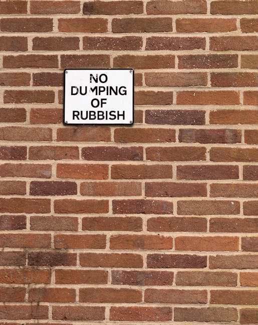 A rectangular white metal sign with black uppercase lettering mounted on a red brick wall. The sign reads 'NO DUMPING OF RUBBISH', with the word 'DUMPING' partially obscured by a black mark. The brick wall has a traditional running bond pattern with varying shades of reddish-brown bricks and light-colored mortar joints, and extends into the background. The sign is positioned approximately in the upper left quadrant of the image, and the lighting is natural, casting soft shadows on the wall. This setting is typical of urban or residential areas where private waste management services such as rubbish collection or on-site clearance are often employed to prevent illegal dumping, aligning with the context of alternative waste handling solutions like those provided by companies similar to <COMPANY_NAME> for rubbish removal near Exhibition Road, South Kensington.