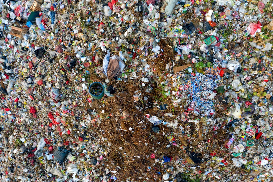 A large pile of mixed waste materials is shown in the foreground, consisting of fragmented and crumpled plastics, metal scraps, and broken electronic components. The waste appears to be discarded rubbish collected from various sources, with textures ranging from smooth, shiny plastic to rough, rusted metal surfaces. The colours in the pile include shades of black, grey, brown, green, and hints of orange and blue, reflecting different materials and contamination. The waste is loosely stacked, creating an uneven, jagged surface that dominates the lower half of the image. In the background, a cloudy sky dominates the upper portion of the image, providing a neutral, overcast atmosphere. The scene appears to be outdoors, possibly on an open site or waste management area, illustrating the type of accumulated rubbish that a professional waste removal service like those by [COMPANY_NAME] might handle in the context of alternative rubbish disposal or on-site clearance, supporting efforts to manage illegal dumping or bulk waste collection near urban areas such as South Kensington.