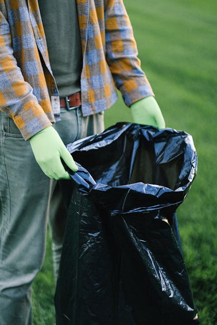 A person wearing a plaid flannel shirt in shades of yellow, orange, and grey, along with green rubber gloves, is holding open a large black plastic rubbish bag outdoors. The bag appears partly filled, positioned in front of the person’s waist, against a background of well-maintained green grass, suggesting a cleanup activity or waste collection effort. The person's grey trousers are visible, secured with a brown belt. The scene is illuminated by natural daylight, casting soft shadows and highlighting the reflective surface of the plastic bag. The setting indicates an environment suitable for private waste disposal or rubbish removal, possibly involving on-site collection of garden or domestic waste. The focus on the disposal activity aligns with the context of alternative waste handling services, like independent rubbish collection, which is often contrasted with council-run refuse collection. The overall composition emphasizes practical waste management, with attention to detail in the materials and environment, supporting professional rubbish removal service descriptions.