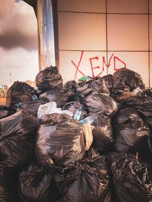 A large accumulation of black plastic rubbish bags is stacked against an exterior building wall made of reflective panels, with some bags leaning against each other and others placed on the ground. The bags appear to be full and tightly sealed, with a few discarded water bottles and miscellaneous waste visible among them. Behind the bags, there is a graffiti message in red spray paint spelling 'XEND' on the wall, indicating an urban environment. The scene is illuminated by natural daylight, with a cloudy sky partially visible in the upper left corner, suggesting this is an outdoor area possibly near a commercial or public space. The setting reflects an instance of unmanaged waste accumulation, highlighting the need for efficient rubbish clearance or alternative waste handling services, such as private collection, provided by companies like rubbishcollectionsouthkensington.co.uk.
