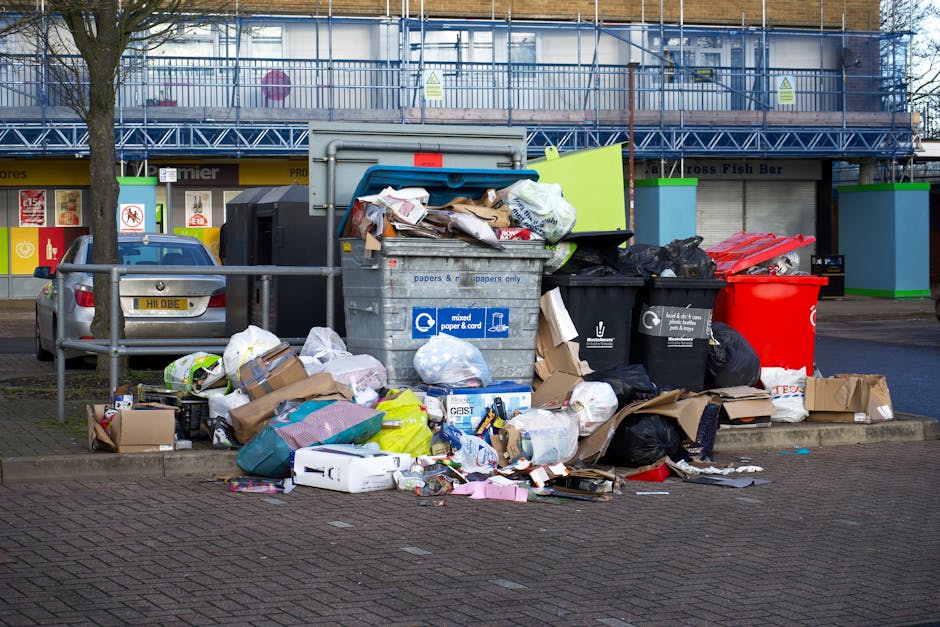 A large collection of overflowing rubbish and recyclable waste bags, cardboard boxes, paper, and plastic packaging are piled on a paved sidewalk area next to a metal railing. The waste is haphazardly stacked around a grey recycling bin labeled for mixed paper and cardboard, with some items spilling onto the ground. Several black and red waste bins are positioned nearby, filled with unsegregated rubbish. In the background, a commercial building with a blue metal staircase and a row of closed shopfronts, including a fish bar, are visible. A silver car is parked adjacent to the waste area, and a leafless tree stands to the left, partially framing the scene. The environment appears to be an urban outdoor setting, with a mix of discarded packaging and waste materials indicating a situation of rubbish piling up, possibly related to private or independent waste collection services, as part of managing refuse outside standard municipal disposal. Natural lighting suggests daytime, and the overall scene is neutral and clear in detail.