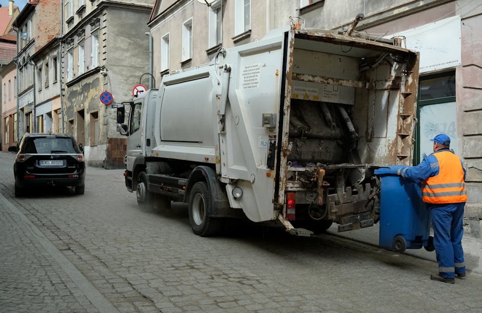 A street scene in an urban area shows a white waste collection truck parked along a cobblestone pavement with its rear hatch open, revealing mechanical parts and an empty cargo area. Nearby, a worker dressed in bright blue workwear and an orange safety vest is placing a blue wheeled rubbish bin into the truck’s rear. The street is lined with multi-story buildings featuring aged, textured facades in shades of beige and gray, with some areas showing signs of peeling paint and weathering. A black car is parked further ahead, and traffic signs indicating parking restrictions are mounted on the building wall. The environment appears calm and typical of a city servicing private waste collection, with natural light illuminating the scene, highlighting the textures of the worn building exteriors, the smooth surface of the truck, and the vibrant safety clothing of the worker. This image reflects a routine rubbish removal operation characteristic of independent waste handling services in urban residential districts.