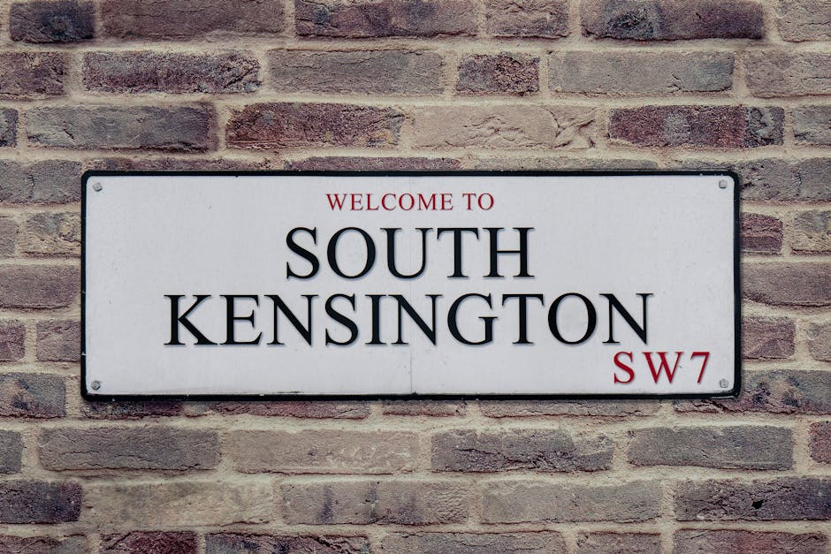 A rectangular metal street sign mounted on a light brown brick wall displaying the text 'WELCOME TO SOUTH KENSINGTON SW7' in black and red lettering. The sign has a white background with black border edges and is attached with four screws, one at each corner. The brick wall behind the sign is composed of uniformly textured bricks with varying shades of light brown and reddish tones, arranged in a horizontal pattern. The scene is evenly lit, with no shadows cast on the sign, and the surroundings suggest an outdoor urban setting, possibly near a residential or commercial property in South Kensington. This image may be relevant in a context where waste removal and independent collection services are discussed, as it represents a typical address identifier within an area serviced by private rubbish clearance providers such as rubbishcollectionsouthkensington.co.uk.
