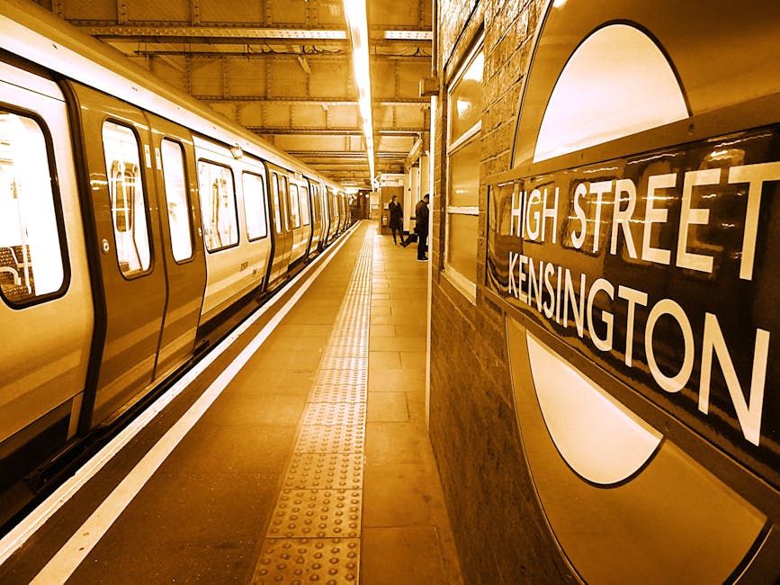 A view of the London Underground station platform at Kensington High Street, with a clean, beige-colored train stopped alongside the platform. The train's exterior features smooth, metallic panels with a glossy finish, and rectangular windows revealing the interior lighting. The platform is illuminated by warm, overhead lighting, and has tactile paving strips running parallel to the train for safety. On the right side of the image, a round London Underground sign with a dark background and white lettering displaying 'High Street Kensington' is mounted on a brick wall, which shows a textured, reddish-brown surface. In the background, several passengers are standing and waiting for the train, some near the edge of the platform, while others are further back, engrossed in their activity. The scene captures an atmosphere of calm urban transit, with crisp details that suggest a well-maintained station environment associated with public transportation infrastructure used by commuters.