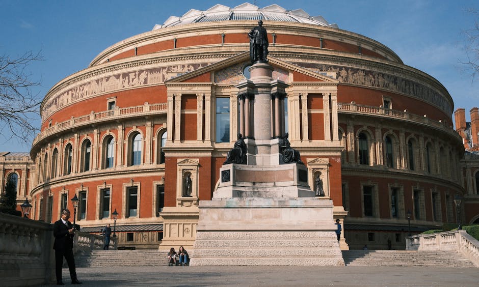 The image features the Royal Albert Hall, a historic circular concert hall with a distinctive domed roof and terracotta-colored brick exterior. The building's façade includes tall, narrow arched windows spaced evenly around its circumference, with decorative stone accents and a prominent stone porter’s lodge at the front. In front of the hall, there is a large stone monument comprising a central pedestal with multiple sculptures and statues, including human figures and ornamental detailing, positioned on a broad, stepped base. The scene is set outdoors on a paved area, with a clear blue sky overhead. A few individuals are visible: a man dressed in dark clothing holding a smartphone on the left side, and two people sitting on the steps near the monument. The environment suggests a typical public gathering space, and the overall view aligns with the context of maintaining clean surroundings in high-profile cultural venues, emphasizing the importance of efficient rubbish removal and dedicated waste handling services by companies like rubbishcollectionsouthkensington.co.uk for event organisers and facility managers.