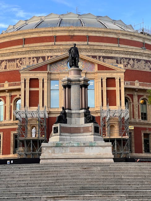 A wide view of the Royal Albert Hall building in South Kensington, showing its round, domed roof with a metallic finish reflecting the daylight. In the foreground, a flight of concrete steps leads up to the entrance, with several people sitting and standing along the sides, some engaged in conversation. A bronze statue of a historical figure on a pedestal is centrally positioned on the steps, with small sculptures or figures inset into the lower part of the pedestal. The hall’s exterior features red brickwork with decorative stone detailing around the windows and along the upper edges of the building. Flanking the staircase are ornate black iron lampposts with spherical lights, and nearby leafless trees and bushes partially obscure adjacent brick buildings. The scene is well-lit with bright, natural sunlight, casting defined shadows and highlighting the historic architectural features, creating a scene that subtly relates to the importance of private waste handling or on-site clearance for large public venues and events such as those held at the Royal Albert Hall, with the emphasis on urban surroundings and the building’s prominent positioning in the historic South Kensington area.