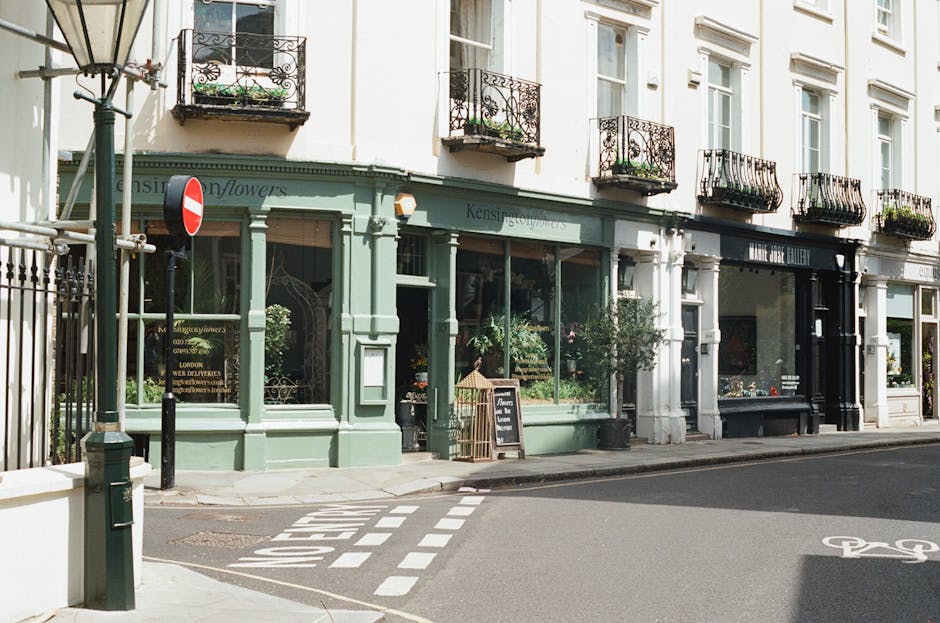 The image depicts a quiet street scene in an urban area, featuring a row of three-story buildings with a combination of commercial and residential facades. The building in the center is painted a soft pastel green color and has large display windows on the ground floor showcasing potted plants and floral arrangements, indicating it may be a florist or a similar shop. The shop has decorative wooden trim around the windows and a small sidewalk chalkboard sign outside. Adjacent to it, there are two other buildings painted in light colors with tall, narrow windows and wrought-iron balcony railings that have flower pots and greenery. To the left, a street lamp with a classic design is visible, alongside a black metal fence and a 'no entry' traffic sign. The pavement features a painted pedestrian crossing, and a bicycle lane is marked nearby on the road surface. The environmental atmosphere is bright with natural daylight, and the scene suggests an area that may handle private waste collection or rubbish disposal outside the commercial properties, aligning with local rubbish removal services.