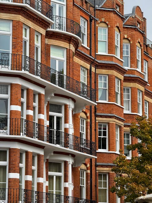 A multi-story red brick residential building with classic Victorian architecture features white decorative trim and black wrought iron railings on the balconies. The building has large sash windows with white frames, some of which are adjacent to small curved bay windows. The balconies are arranged in a repetitive pattern across the front façade, each supported by white columns and separated by horizontal white bands. In the lower right of the image, a tree with sparse yellowish-green leaves partially obscures the view, suggesting an outdoor urban environment. The overall scene exhibits clean, well-maintained external brickwork with a mixture of warm red tones and light-colored accents, indicative of a traditional London-style building. The atmosphere appears natural daylight, highlighting the architectural details and textures of the bricks and railings, consistent with a residential area where private waste collection services might operate for such properties.