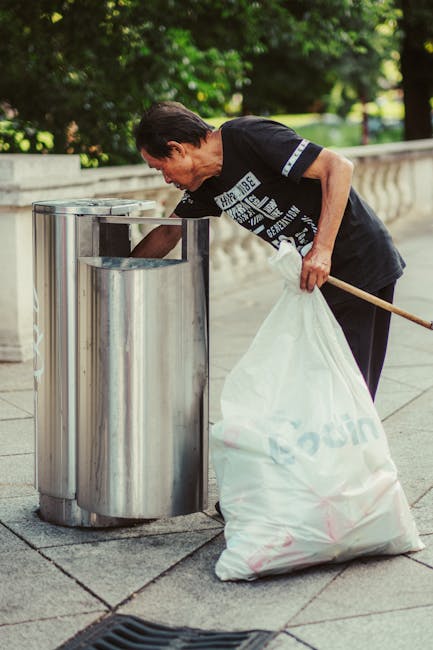A man with dark hair, wearing a black T-shirt with white printed text and dark trousers, is bending over as he disposes of rubbish into a tall, cylindrical stainless steel public litter bin on a paved sidewalk. The bin is split into multiple sections with open tops, designed for waste separation, and has a polished finish that reflects light. He holds a large, white plastic bag filled with waste in his left hand, which rests against his body, and uses his right hand to reach inside the bin. The environment around him features lush green trees and bushes in the background, suggesting a park or garden nearby. The scene appears during daylight with soft natural lighting, emphasizing the clean, orderly setting typical for urban waste disposal or private rubbish collection. This image illustrates the concept of independent rubbish disposal, aligning with private waste management services that focus on efficient refuse collection and street cleaning.