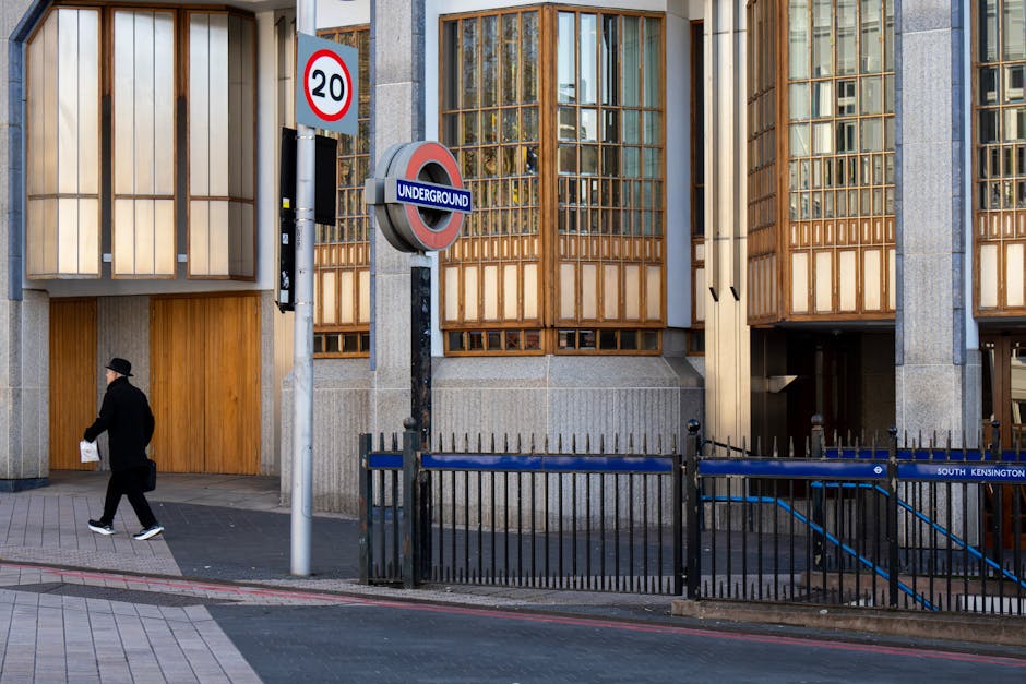 A street scene showing the entrance to an underground transit station with a prominent roundel sign reading 'UNDERGROUND' mounted on a weathered black pole. The station entrance is adjacent to a modern building façade featuring large windows framed with wooden paneling, some of which are partially open or divided by vertical wooden slats. To the left, a man dressed in dark clothing, including a black hat, is walking along the pavement carrying a small white bag. In the foreground, a black metal fence runs parallel to the curb, separating the sidewalk from the street. A red and white circular speed limit sign displaying '20' is positioned above a rectangular electronic sign affixed to the pole, indicating traffic regulations. The roadway surface is smooth with a red-painted curb and a light grey tiled sidewalk. The environment appears clean and well-maintained, reflecting typical urban infrastructure associated with public transit access points, which may be relevant in the context of alternative waste handling or site clearance services like those offered by rubbishcollectionsouthkensington.co.uk in the South Kensington area.